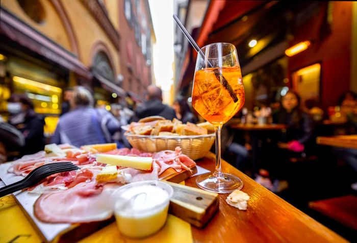 A delightful outdoor table setting with thinly sliced cured ham, cheese, a basket of fresh bread, and a refreshing drink.