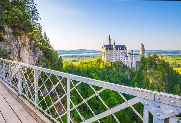 The stunning Romanesque Revival architecture of the iconic Neuschwanstein Castle is enveloped by lush forests, as seen from the nearby bridge.