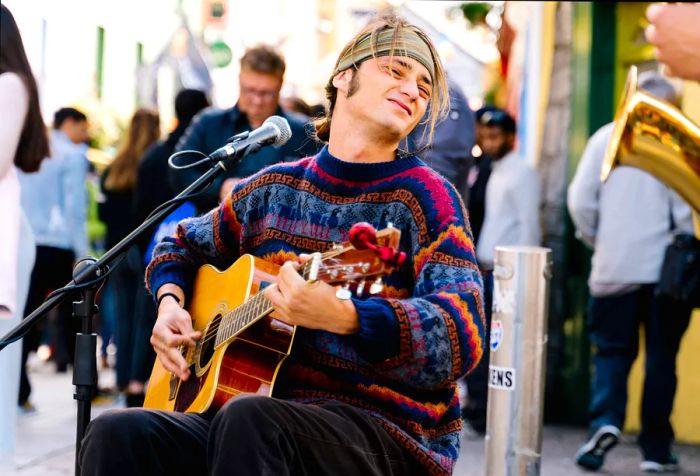 A gifted musician performs on the street, strumming a guitar by a microphone, dressed in a colorful cardigan and bandana, enchanting passersby with heartfelt melodies.