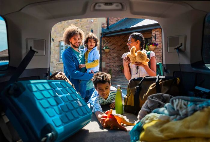 A family of four is unloading their bags and children's toys from the back of their car.