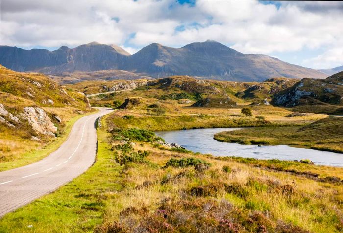 A road meanders alongside a river and gentle hills, offering distant glimpses of a mountain range.