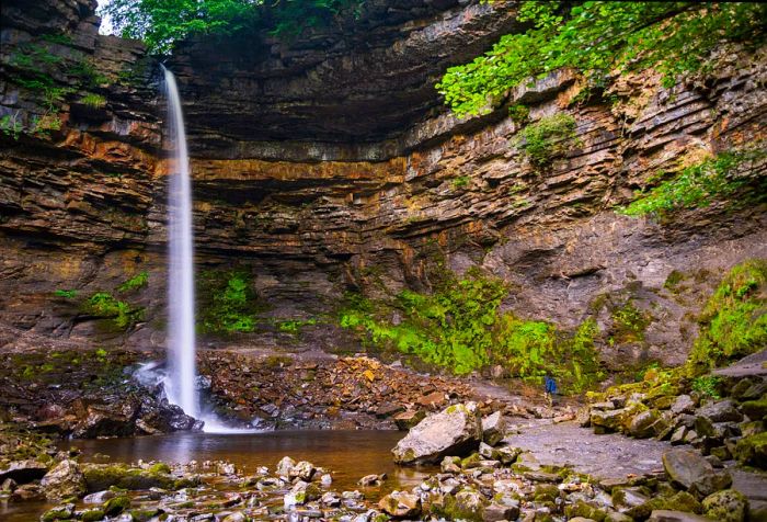 A magnificent waterfall descends directly to the ground below, encircled by rocky cliffs, stones, and a few plants.