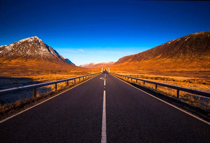 A two-lane asphalt road winds along a cliff on one side, with a cone-shaped mountain on the other, leading toward snow-capped ranges in the distance.