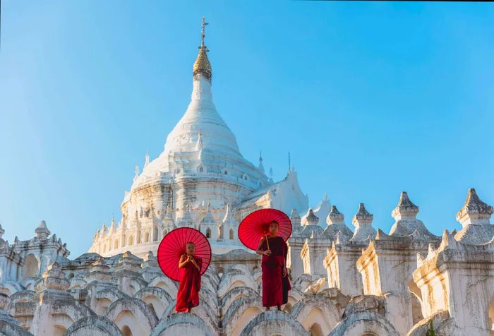 Two young Buddhist monks with red umbrellas stand atop the undulating layers of a large white pagoda.