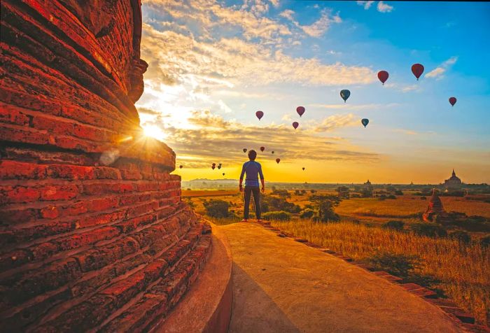 A man stands next to a crumbling temple wall, gazing up at hot air balloons drifting against a picturesque sky.