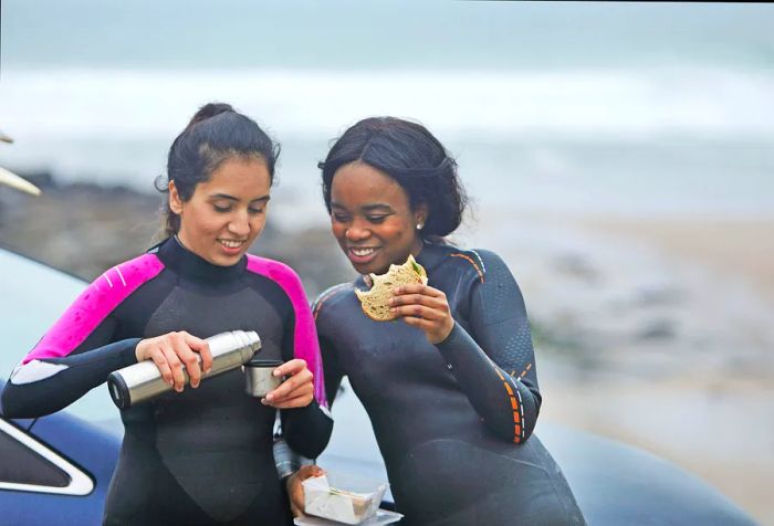 A group of friends in wetsuits enjoying refreshments and a sandwich by their car.