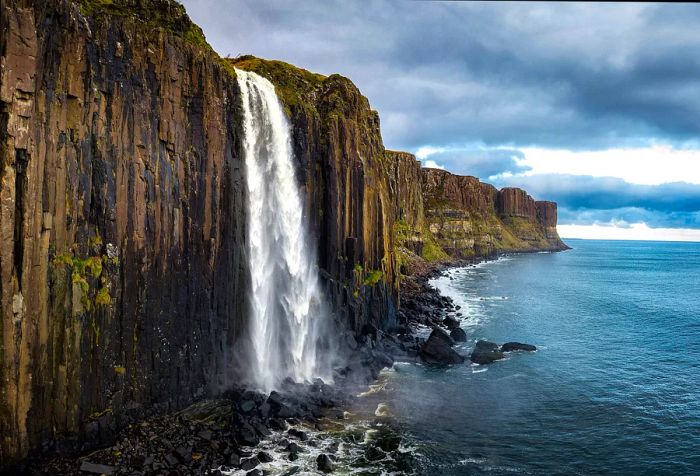 An incredible waterfall cascades into the sea from a cliff, surrounded by a dramatic formation of vertical basalt columns.