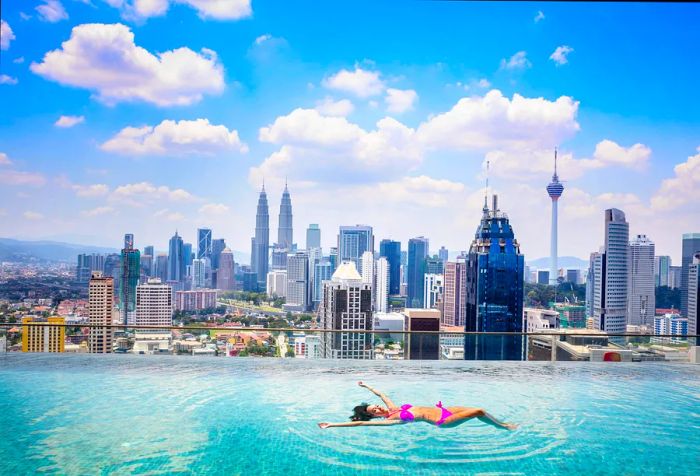 A woman in pink swimwear relaxes in an infinity pool, overlooking modern skyscrapers beneath a cloudy blue sky.