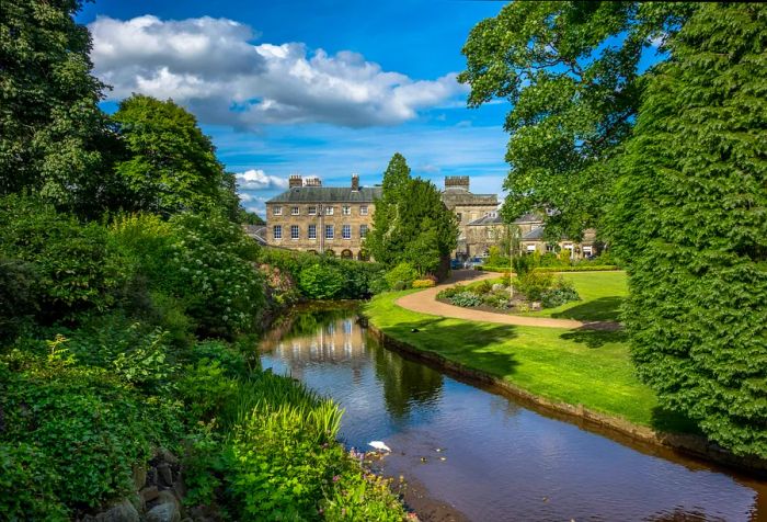 At the end of a grassy path lined with plants and trees, an ancient stone building stands beside a tranquil pond.