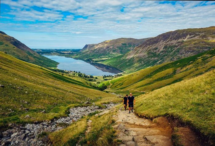 Two people climbing a stone staircase between two green valleys, with a lake visible in the distance.