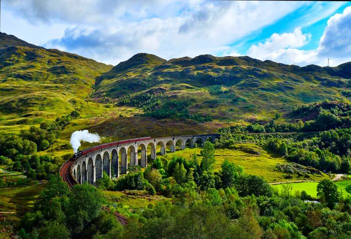 A steam train crosses a viaduct amidst a mountainous landscape blanketed in lush greenery.