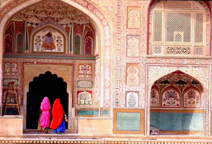 Two women elegantly draped in vibrant linen stroll through a pointed archway, flanked by walls embellished with intricate carvings.