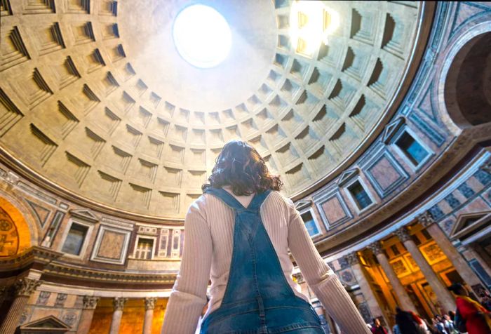 A woman stands inside a building featuring a coffered dome, light streaming through the central oculus above.