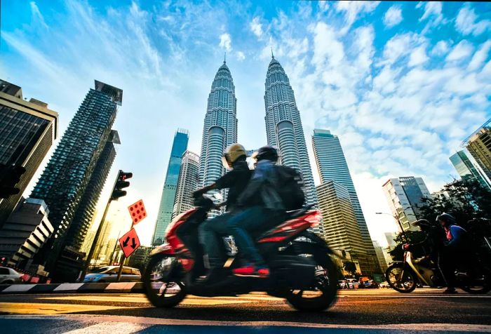 Cars navigate a vibrant street lined with the iconic Petronas Twin Towers amidst towering buildings.