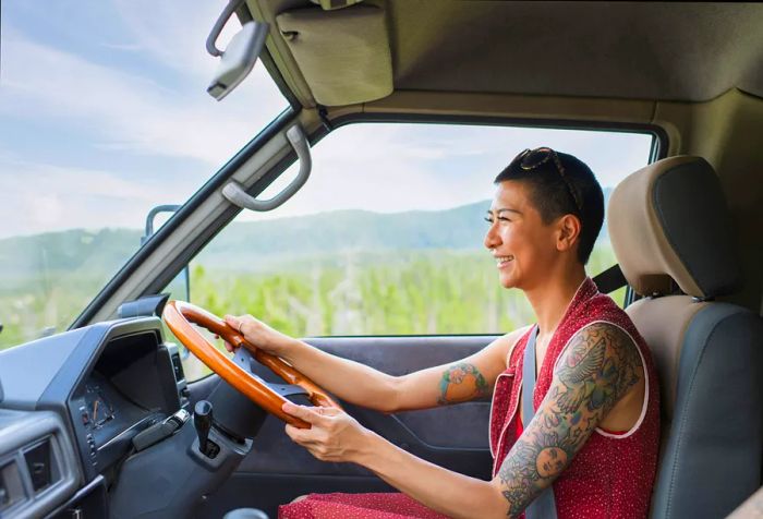 A cheerful woman with tattoos and a shaved hairstyle, dressed in a red dress, is driving a car.