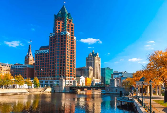 Downtown Milwaukee's skyline, prominently featuring Milwaukee Center, with the Milwaukee River and vibrant orange trees in the foreground during autumn.