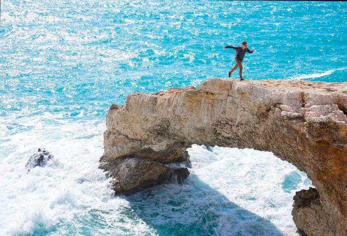 A runner navigates the top of a stunning arch rock formation as waves crash below in the ocean.