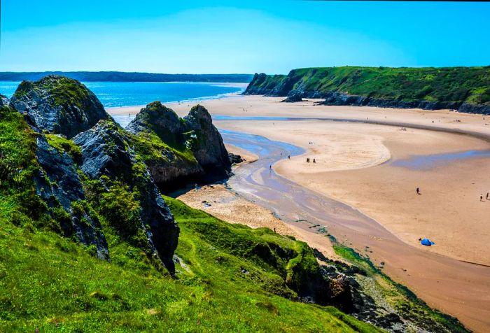 A sprawling sandy beach nestled between rugged, moss-covered cliffs, with a stream meandering through the golden sand into the ocean.