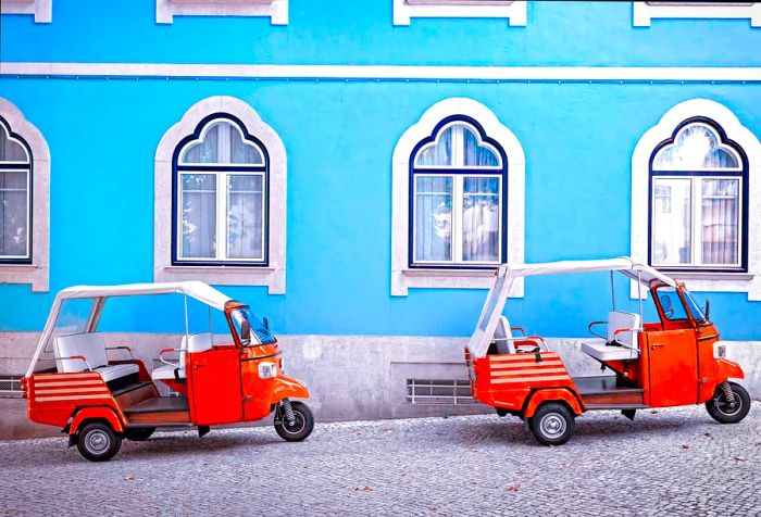 Two orange tuk-tuks are parked along a cobblestone street next to a building painted blue.