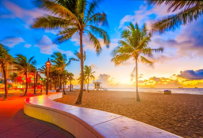 A lifeguard tower and palm trees rise prominently on the deserted sandy beach next to a promenade, all bathed in a picturesque sunset.