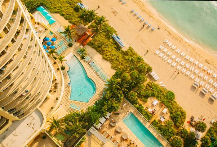 An aerial shot of a resort featuring tall residential buildings overlooking swimming pools that lead to the beach.