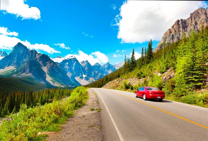 A solitary red car heading toward majestic rock mountains with sharp peaks on a bright, sunny day.