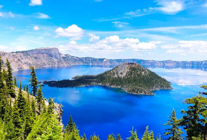 A pristine lake with deep blue waters, surrounded by mountains and featuring a hill adorned with lush green trees in the center.