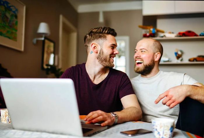 A cheerful gay couple is sitting at a dining table, working on a laptop together.