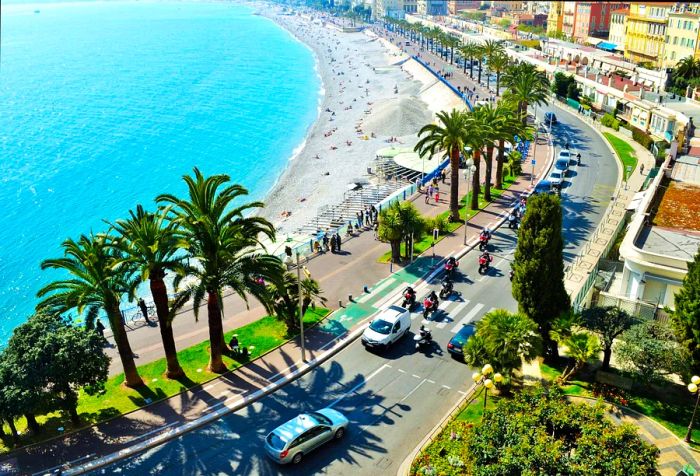 A bustling beach adjacent to a tree-lined promenade and roadway.