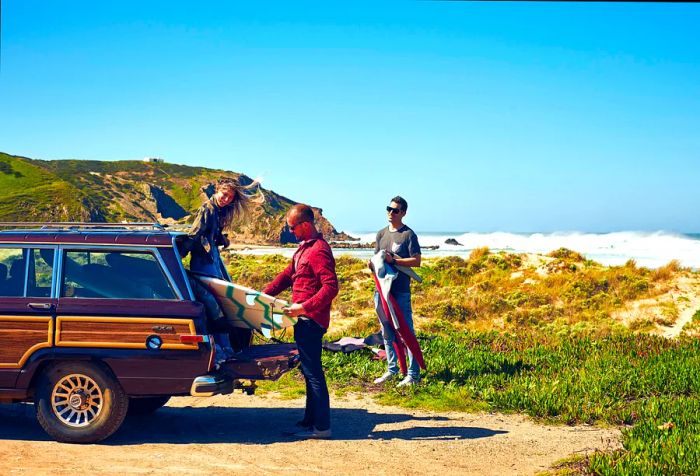 Three friends are unloading their surfboards from the trunk of a car parked on a grassy beach.