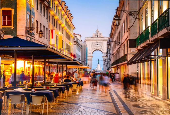 Tourists stroll along a cobblestone street lined with outdoor dining, heading toward an archway between buildings.