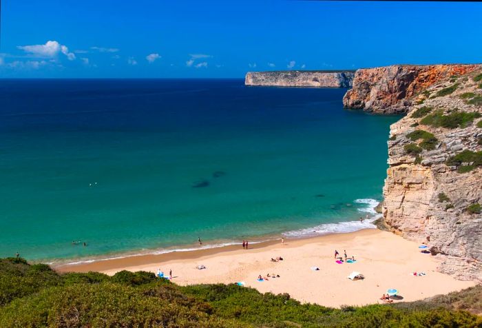 A sandy beach stretches alongside the calm blue sea, framed by towering cliffs beneath a clear blue sky.