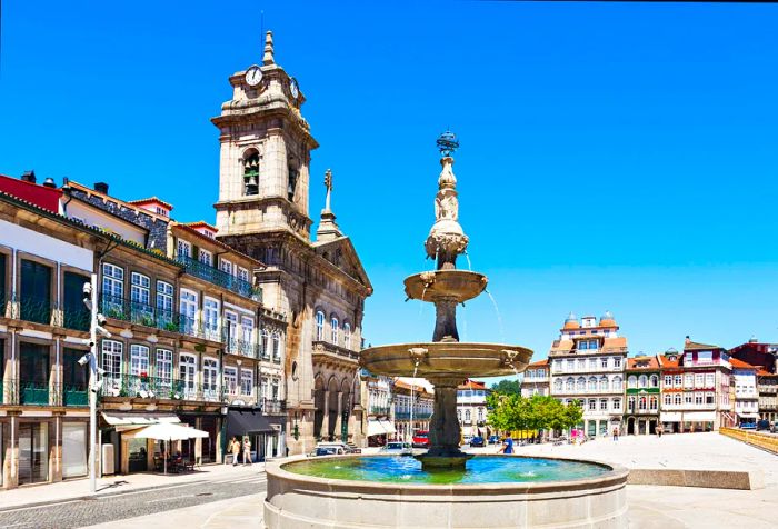 A fountain graces a church square, surrounded by beautiful historic architecture.