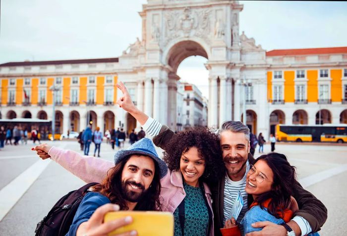 Four friends happily capture a group photo with a smartphone in a public square featuring an impressive arch monument in the background.