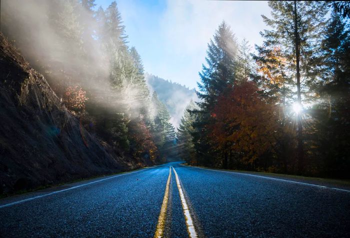 A two-lane road runs alongside a rocky mountain, with foggy autumn trees in the background and bright sunlight shining down.