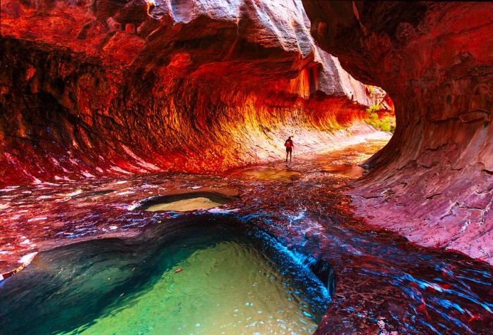 An individual strolls through a narrow canyon resembling a subway tunnel, complete with a swimming hole.