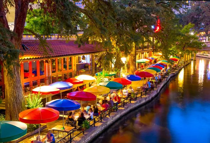 A picturesque riverwalk comes alive with colorful parasols and charming eateries, all framed by majestic trees.