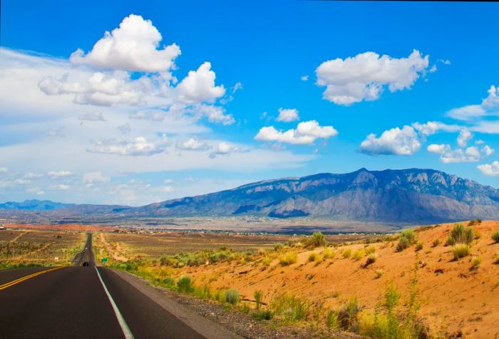 A long, paved highway stretches across a desolate landscape, with a stunning mountain range silhouetted against the cloudy blue sky.