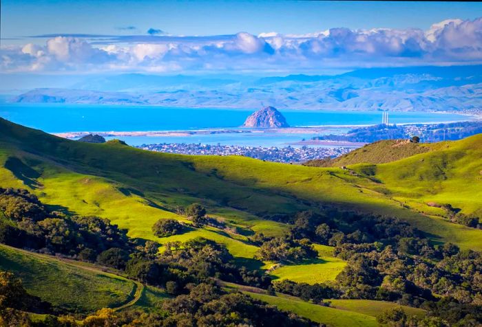 A towering rock sits in the middle of a bay near a coastal village, viewed from sloping green hills.