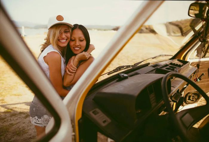 Two joyful women embrace in front of a vehicle parked by the seaside.