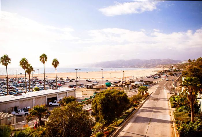 A bustling highway runs alongside a wide sandy beach next to a parking lot that boasts an expansive view of the ocean.