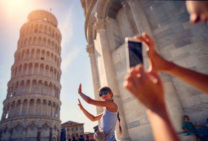 A phone captures a moment as a young woman poses with her arms raised beside the iconic Leaning Tower of Pisa.