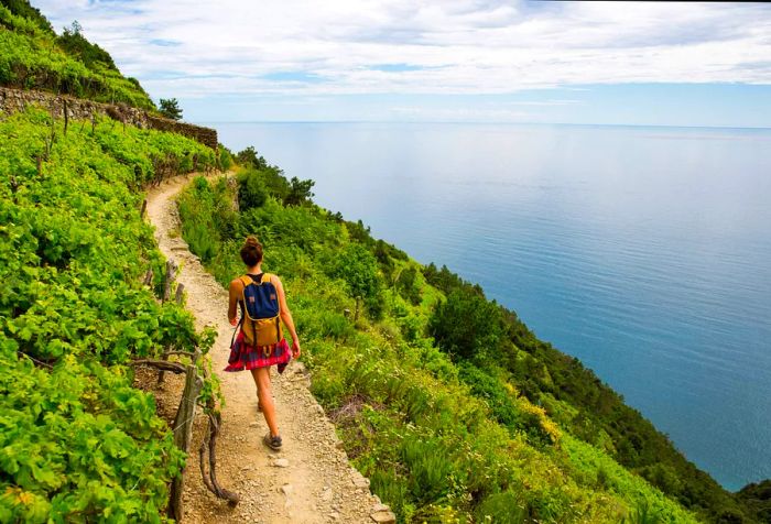 A traveler with a yellow backpack strolls along a narrow sandy path on a mountain blanketed in lush greenery, above the shimmering blue sea.