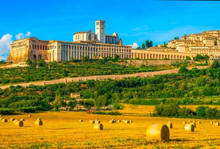 The stunning Basilica of Saint Francis rises above the town of Assisi, which overlooks a field dotted with hay bales.