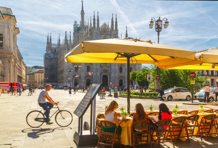 Three friends enjoy a meal outdoors beneath an umbrella in a bustling square near a church.