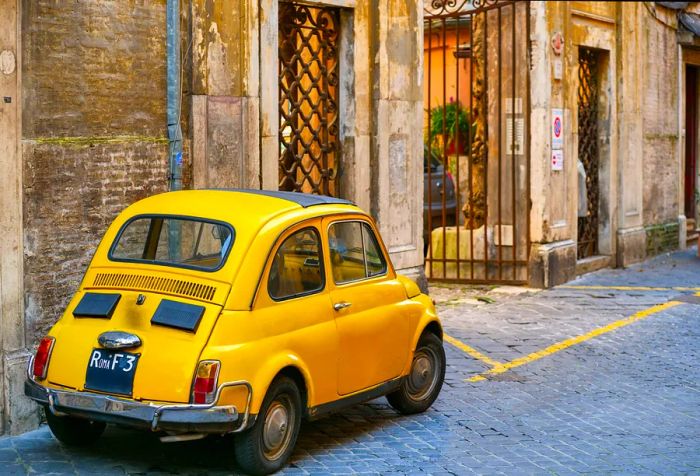 A classic yellow vintage car sits parked beside a gate in a charming alleyway.