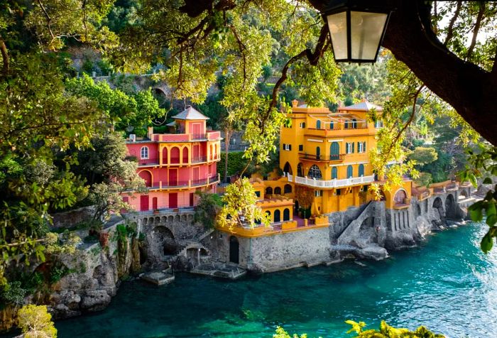 A vibrant yellow and red villa perched on the edge of a cliff, surrounded by tree branches overlooking the coast.