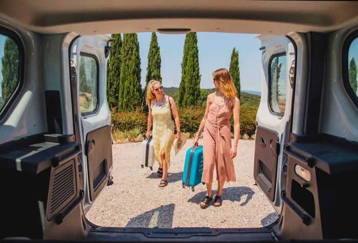 Two women exchange glances as they load their luggage into the trunk of a car.