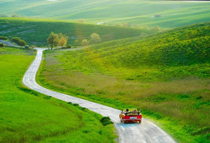 A couple drives a red car down a dusty white road, heading towards lush green fields.