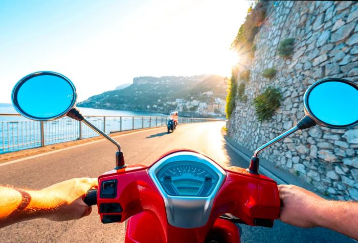 Two drivers navigate in opposite directions on a road bordered by a rocky wall next to the harbor.